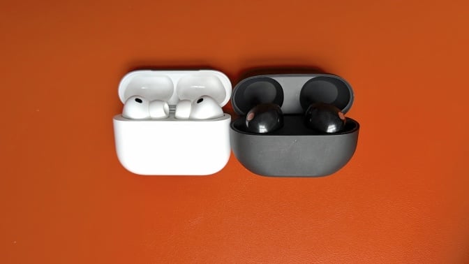 A photo of the Apple AirPods Pro 3 sitting on an orange table next to the Sony WF-1000XM5, both in their case